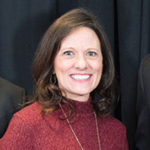 A woman with long brown hair, wearing a maroon sweater and a necklace, smiles in front of a black background.