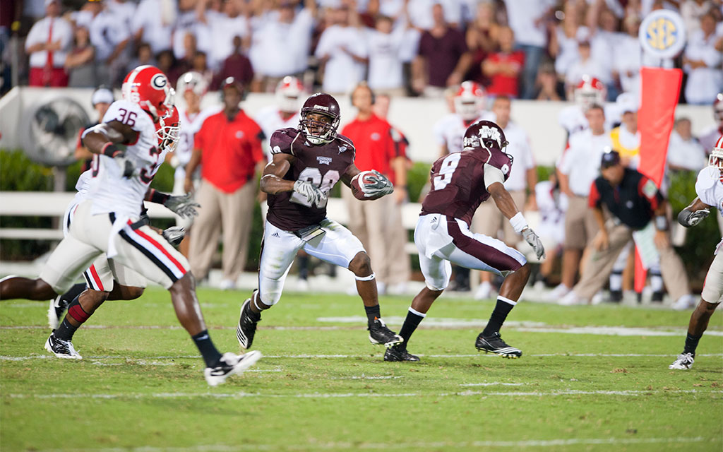 A college football player in a maroon uniform runs with the ball while opponents in red and white uniforms attempt to intercept him on the field.