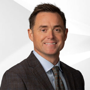 Man in a suit and tie smiling at the camera against a plain, light-colored background.