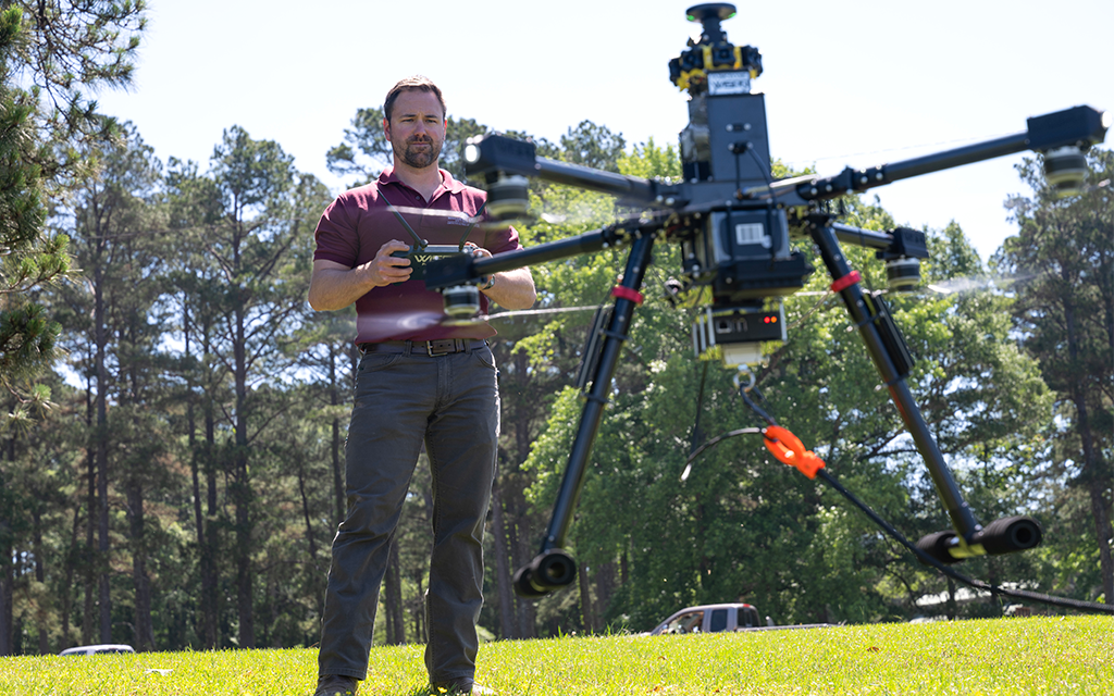 A man operates a large drone outdoors on a grassy area with trees and cars in the background.