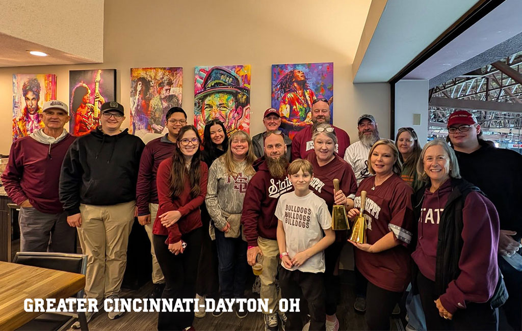 A group of people wearing maroon and gray clothing pose together indoors in front of colorful artwork. Text at the bottom reads “Greater Cincinnati-Dayton, OH.”.