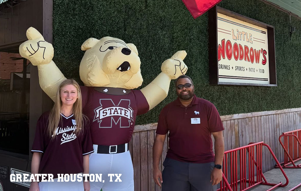 Two people stand beside a large inflatable bulldog mascot wearing a Mississippi State shirt outside Little Woodrow’s in Greater Houston, TX.