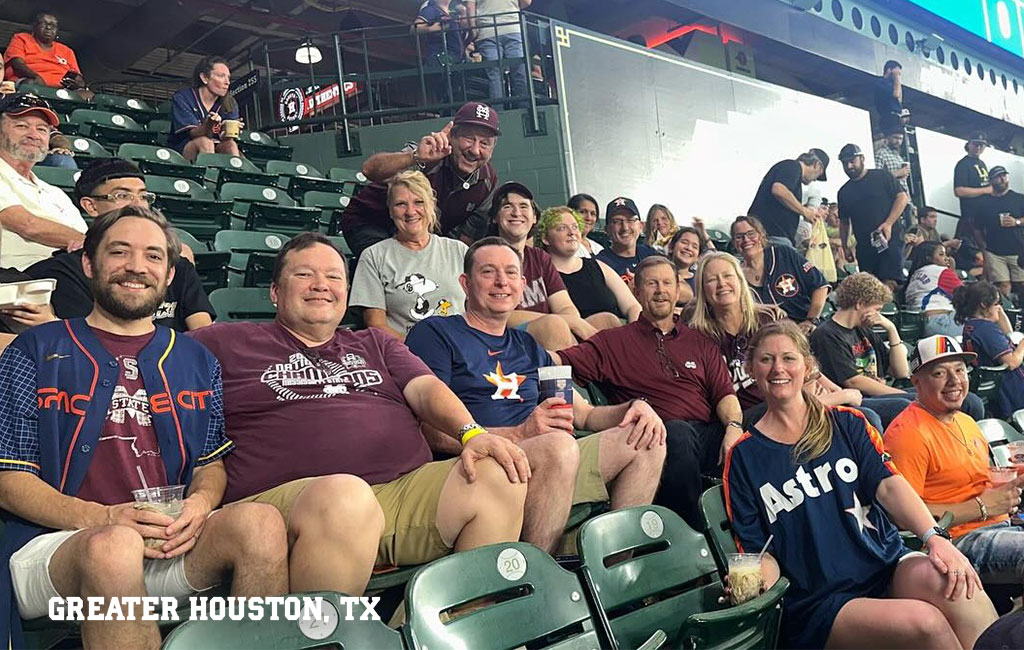 A group of people wearing Astros and Aggies shirts sit together in stadium seats, smiling at the camera. Text reads “Greater Houston, TX” in the corner.