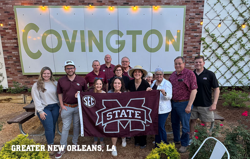 A group of thirteen people pose outdoors in front of a “Covington” sign, holding a Mississippi State University flag. Text reads “Greater New Orleans, LA” in the lower left.