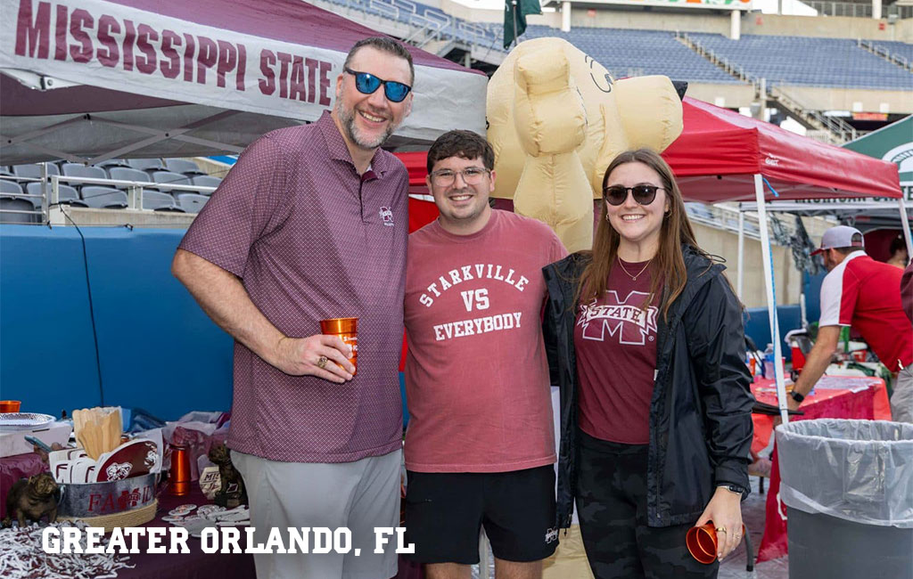 Three people in Mississippi State apparel stand and smile at an outdoor event with tents and tables. A large inflatable object is in the background. Text reads “GREATER ORLANDO, FL”.