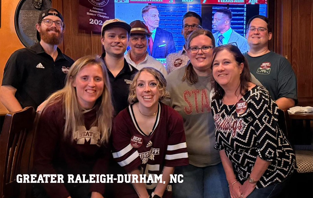A group of people in Mississippi State attire pose together in a restaurant; a TV behind them shows sports commentators; text reads “GREATER RALEIGH-DURHAM, NC.”.