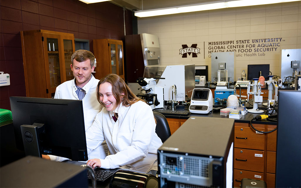 Two researchers in lab coats work together at a computer in a laboratory at Mississippi State University’s Global Center for Aquatic Health and Food Security.