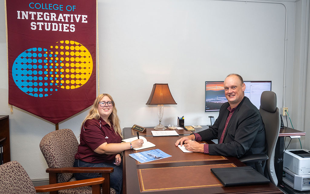 Two people sit across from each other at a desk, with paperwork in front of them, in an office. A banner reads “College of Integrative Studies.”.