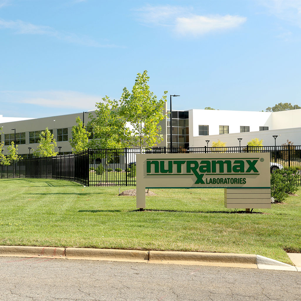 A building with large windows and trees in front, behind a black metal fence. A sign in the grass reads “Nutramax Laboratories” in green letters.