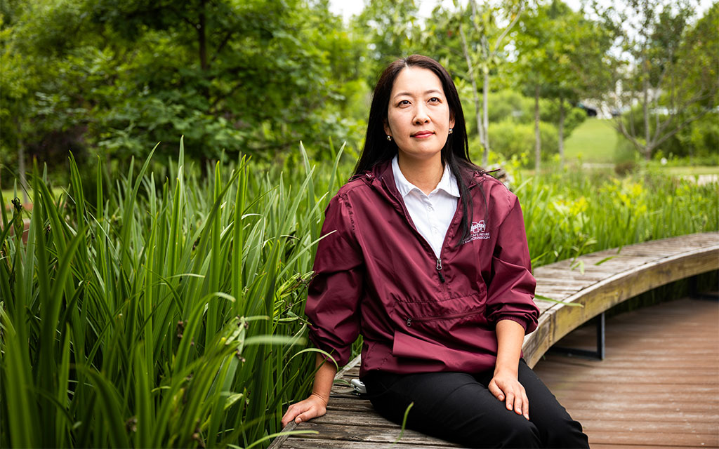 A woman in a maroon jacket sits on a curved wooden bench surrounded by tall green plants and trees in a park.