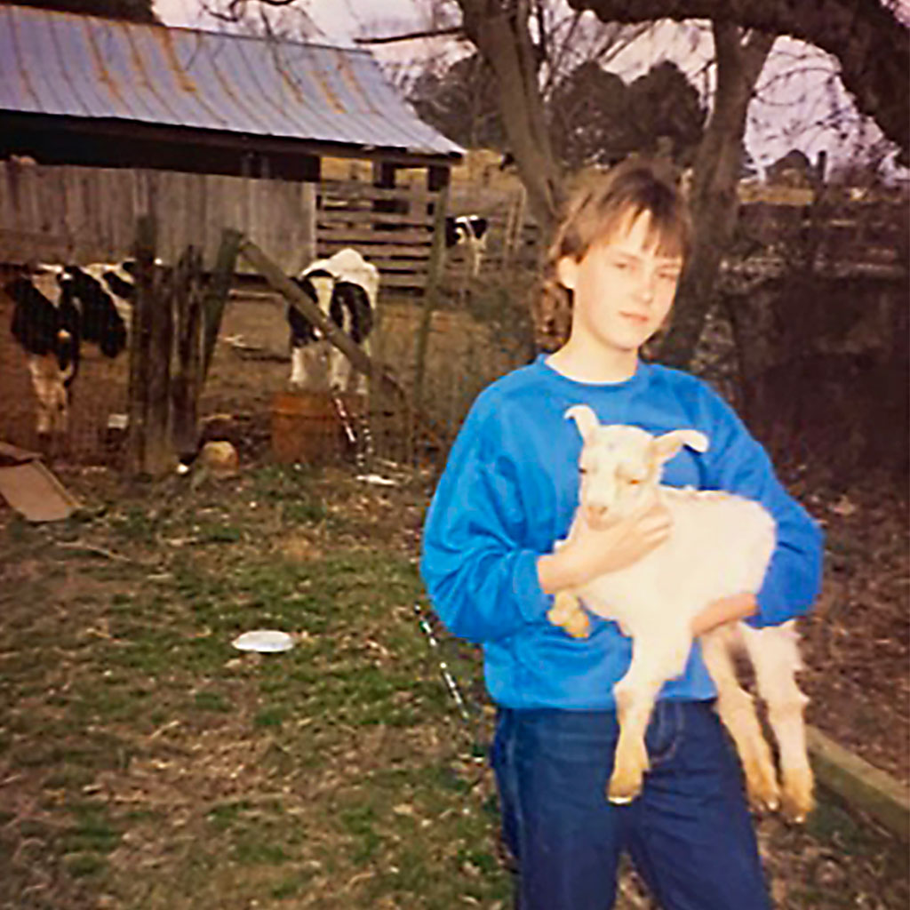 A person in a blue sweatshirt stands outside, holding a white goat. Cows and a wooden shed are visible in the background on a farm.