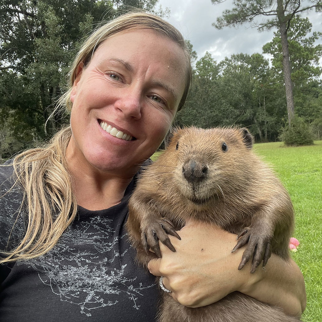 Woman smiling and holding a beaver outdoors, with trees and grass visible in the background.