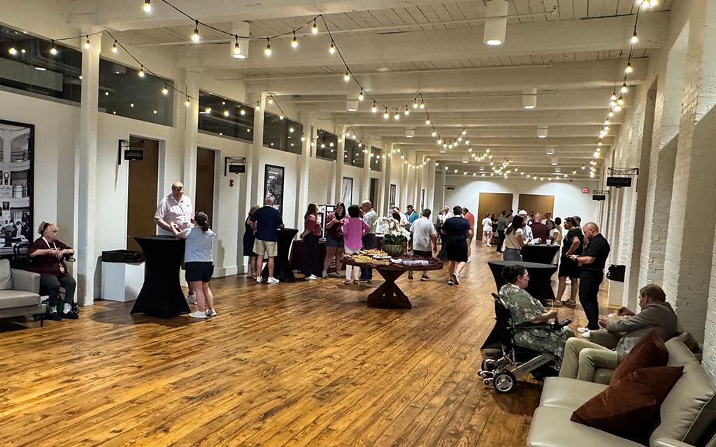 People gather in a well-lit hallway with wooden floors, string lights, and refreshments on tables. Some are seated, while others stand and converse.