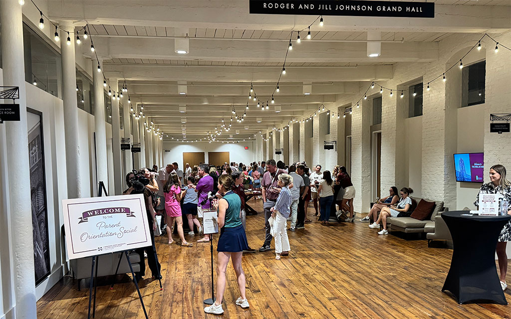 A group of people gather in a large, bright hallway with white walls and wooden floors for a “Parent Orientation Social” event. A welcome sign is visible in the foreground.