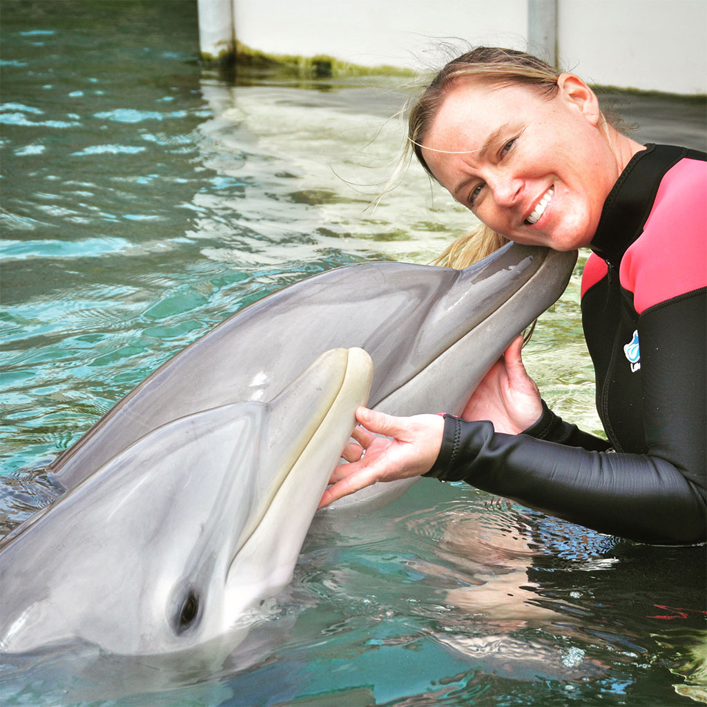 A woman in a wetsuit smiles while holding a dolphin’s snout in a pool.