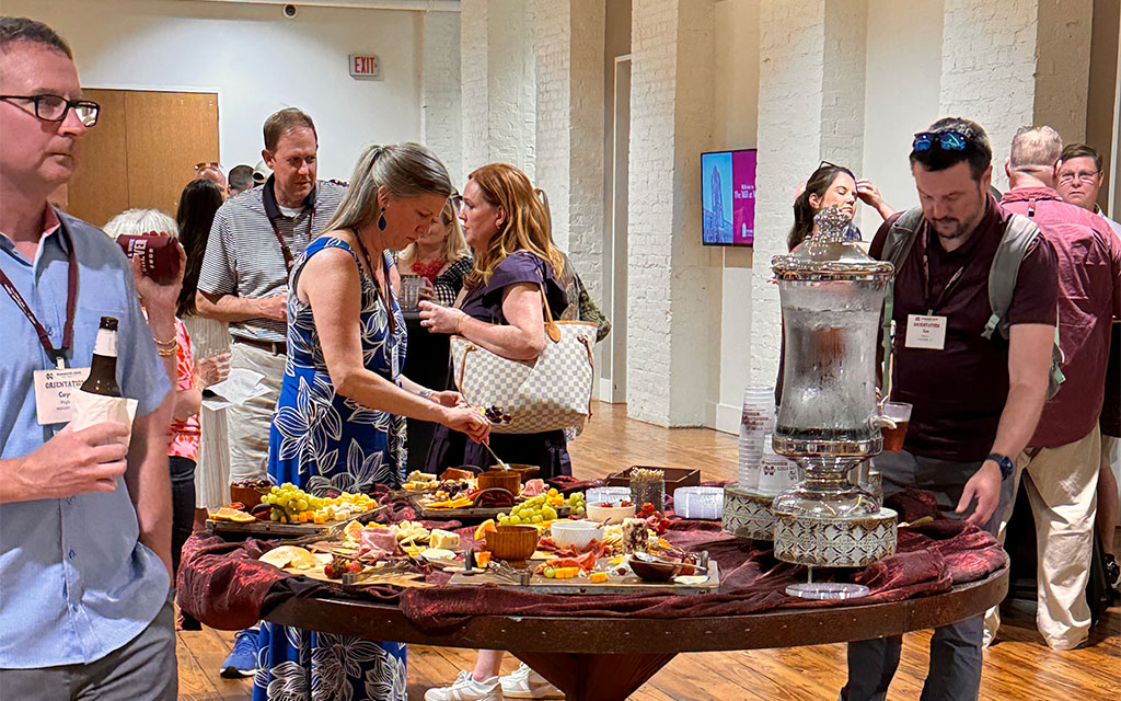 People gather around a table with assorted snacks and a large beverage dispenser at an indoor social event.