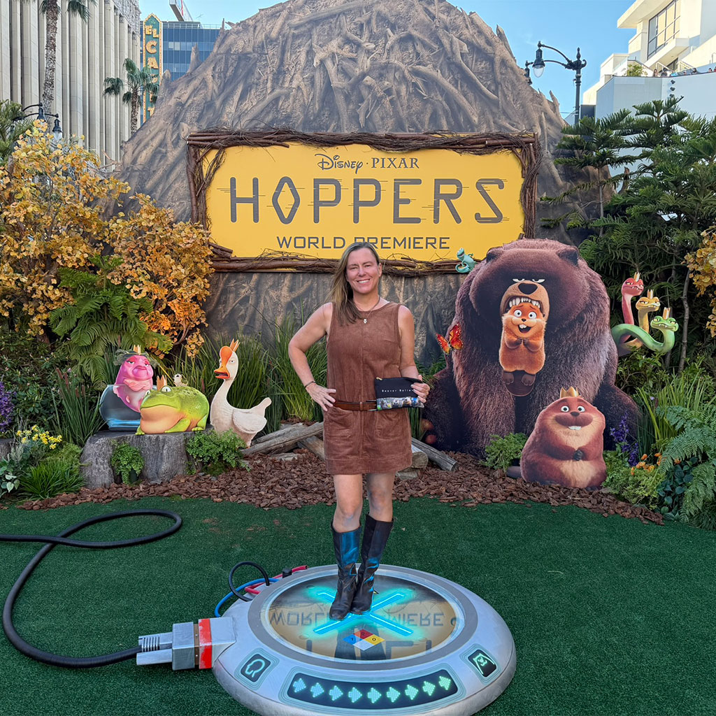 A woman stands on a circular platform in front of a “Disney Pixar Hoppers World Premiere” backdrop with animated animal characters and greenery.