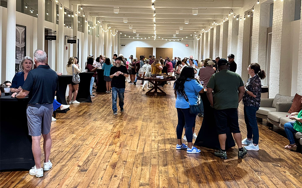 People gather and socialize in a spacious, well-lit hall with wooden floors and white walls. Some stand in groups while others walk through the area.