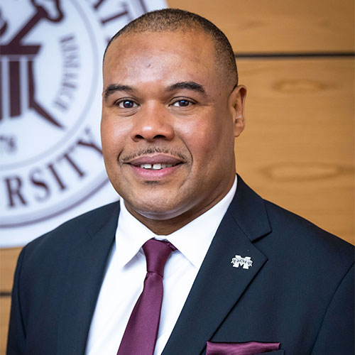 A man in a suit and tie smiles in front of a university seal on a wood-paneled wall.