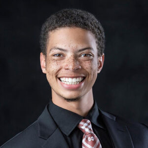 Young man with short curly hair and freckles, wearing a black suit, black shirt, and striped tie, smiling at the camera against a dark background.