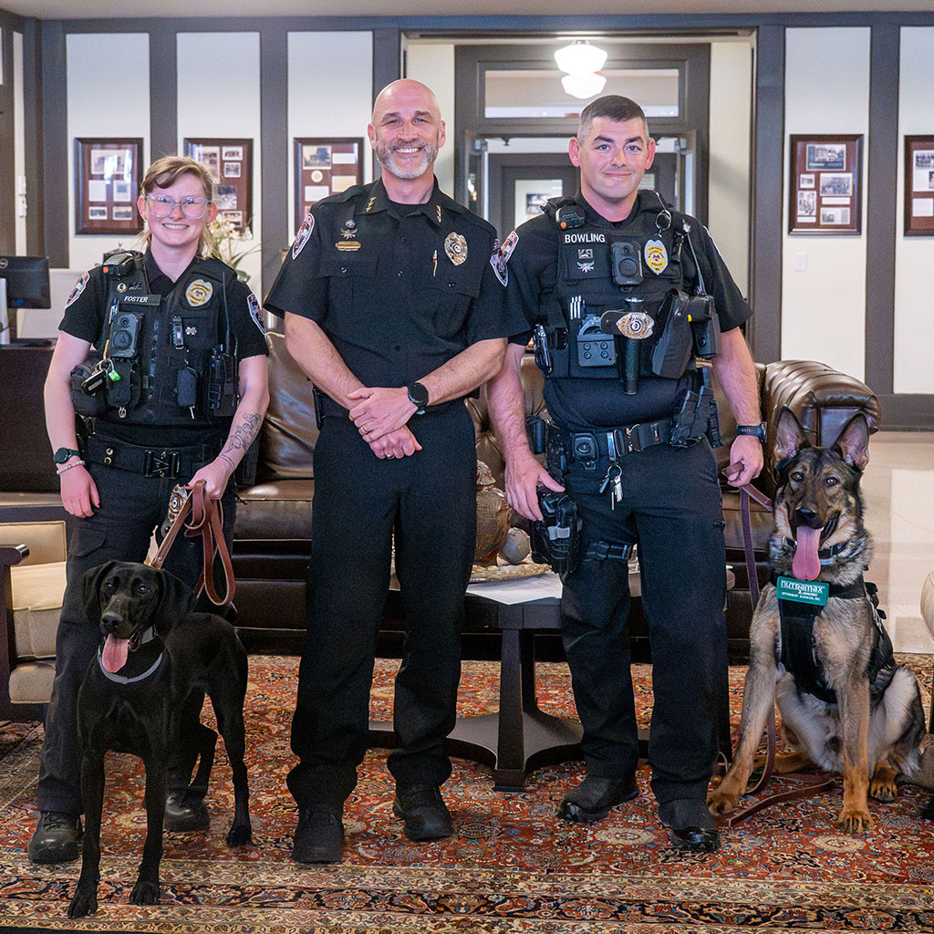 Three police officers and two dogs stand together in a lobby area, posing for a group photo on a patterned rug with couches and framed pictures in the background.