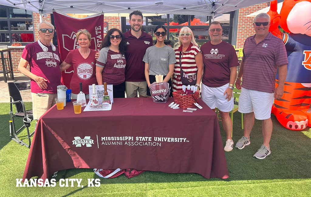 Eight people stand behind a Mississippi State University Alumni Association table under a canopy at an outdoor event in Kansas City, KS. Most are wearing maroon MSU shirts.