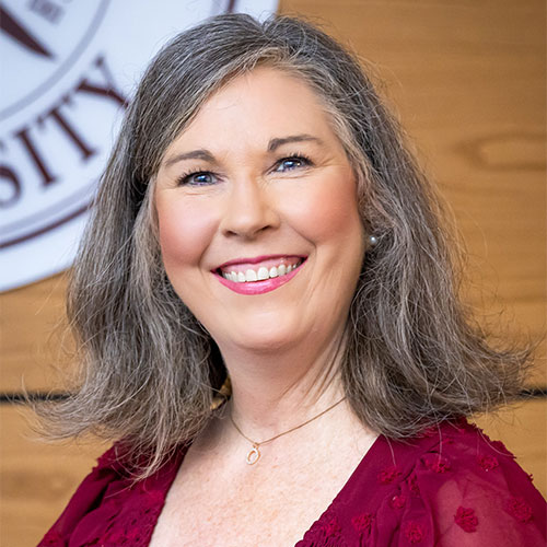 Smiling woman with shoulder-length gray hair wearing a burgundy blouse, standing indoors in front of a partial university sign.