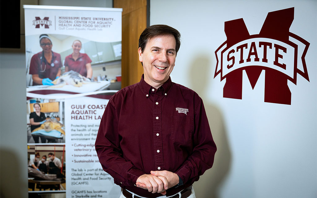 A man in a maroon Mississippi State University shirt stands indoors beside a university-branded banner and logo, smiling at the camera.