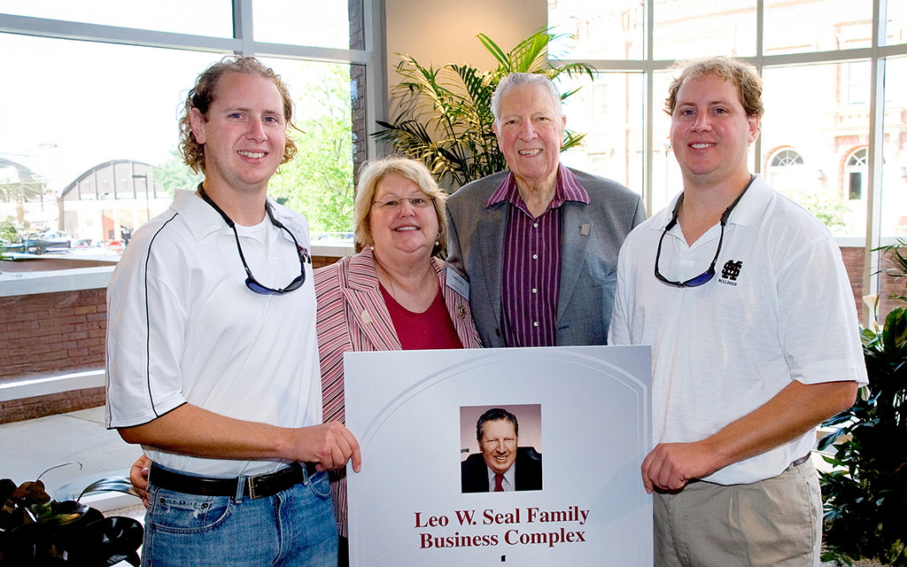 Four people stand indoors holding a sign that reads “Leo W. Seal Family Business Complex” with a photo of an older man, large windows and plants in the background.