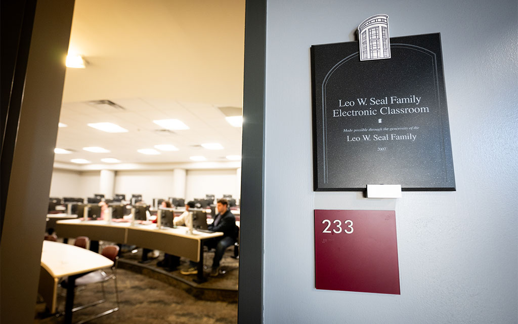 Open classroom door reveals students seated at computers; sign outside reads “Leo W. Seal Family Electronic Classroom,” with room number 233 visible below.
