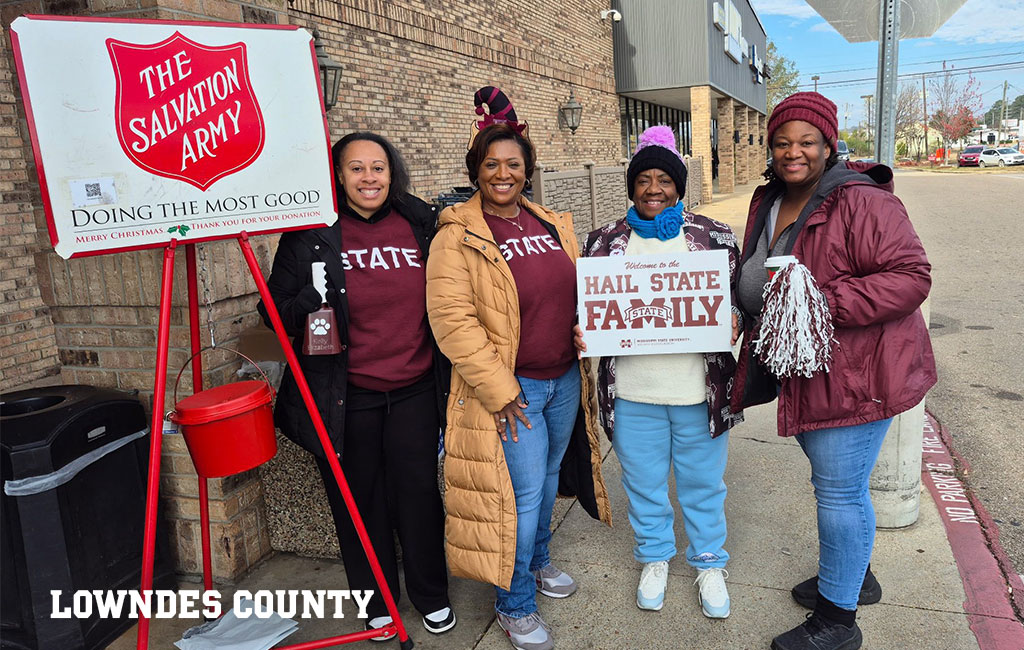 Four women stand by a Salvation Army donation kettle on a sidewalk, three wearing “STATE” shirts or jackets, one holding a “Hail State Family” sign. Building and street visible in background.