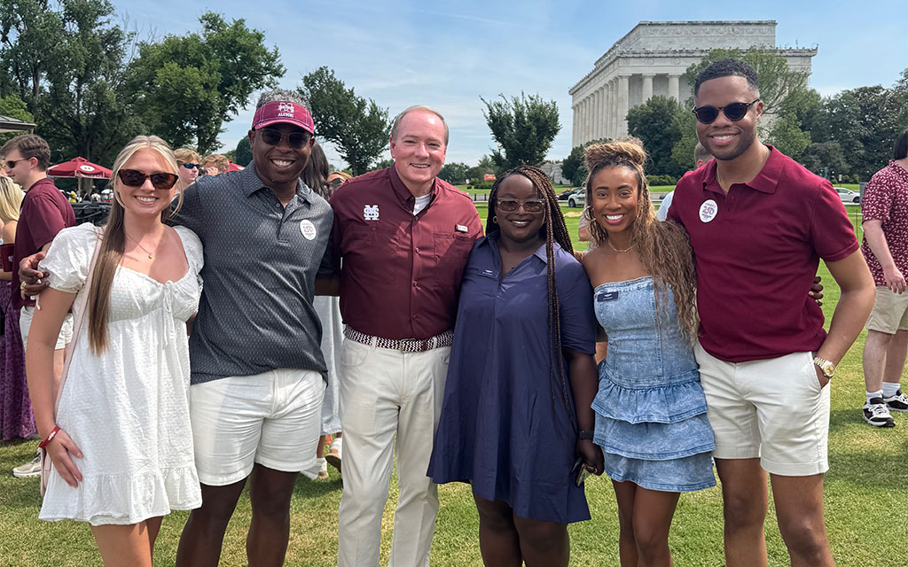 Six people stand together smiling outdoors on grass with trees and the Lincoln Memorial in the background on a sunny day.