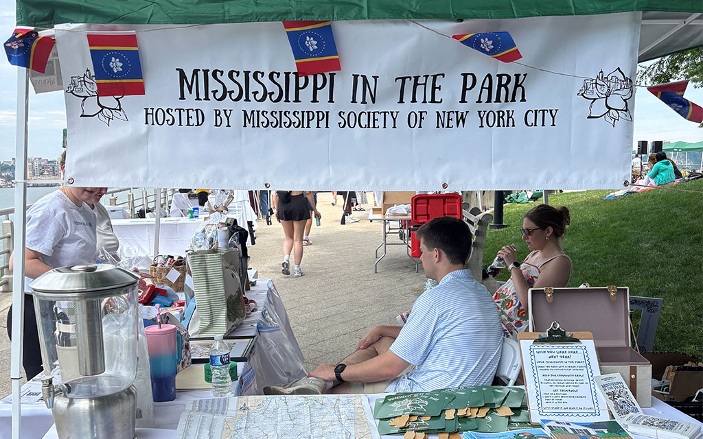 A booth with a “Mississippi in the Park” banner, hosted by Mississippi Society of New York City, displays maps and items; people are seated and a river is visible in the background.