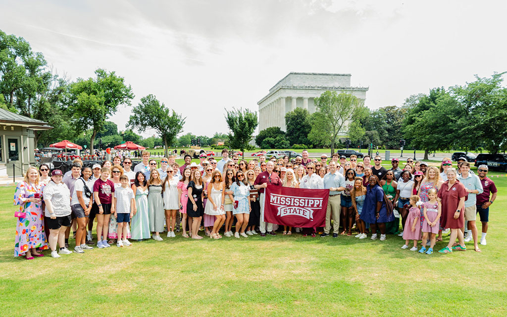 A large group of people poses on grass holding a “Mississippi State” banner with the Lincoln Memorial visible in the background.
