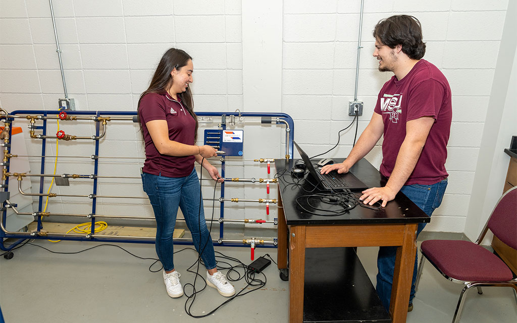 Two people operate lab equipment and a computer in a classroom, with pipes and sensors connected to a control panel on the wall.
