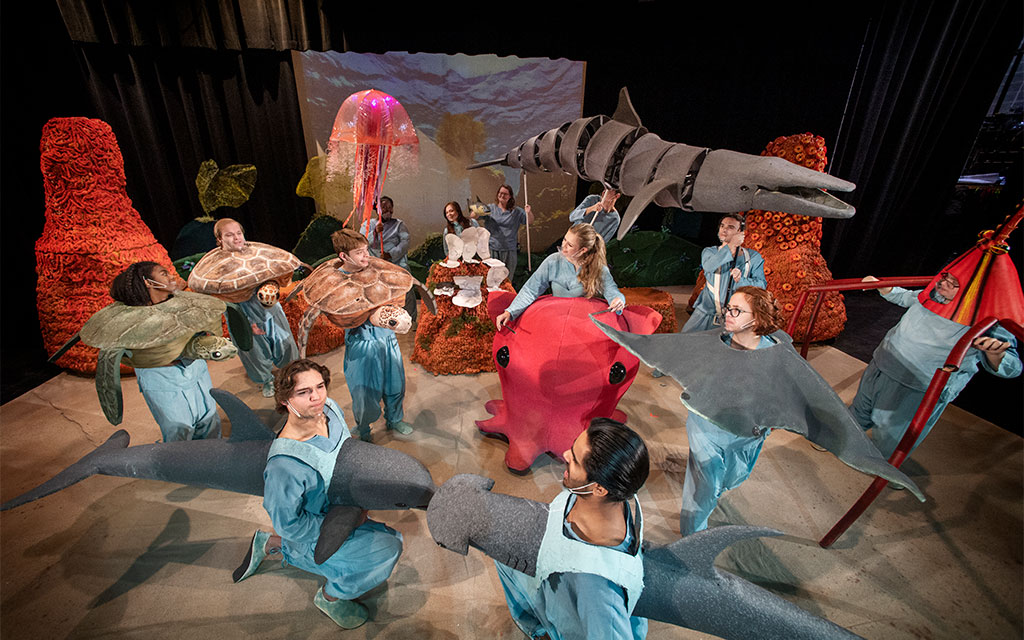 People in costumes resembling sea creatures perform on stage with ocean-themed props and backdrop.