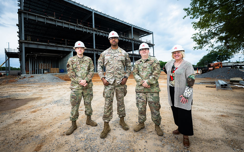 Four people wearing hard hats, three in military uniforms and one in civilian clothing, stand in front of a large building under construction at a dirt-covered site.