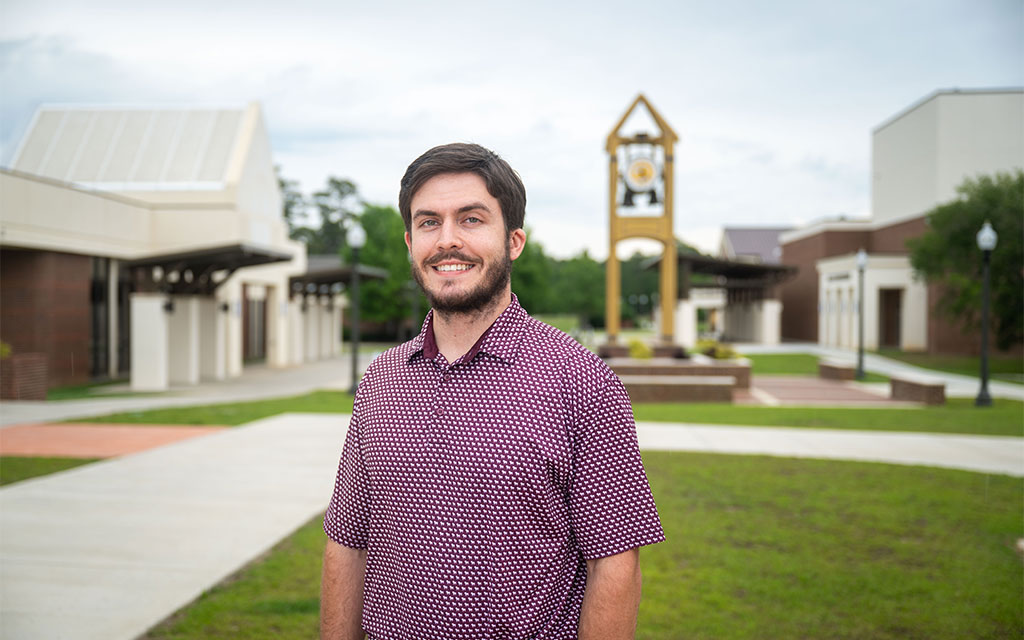 A man with brown hair and a beard stands outside on a campus, smiling at the camera, with a clock tower and academic buildings in the background.