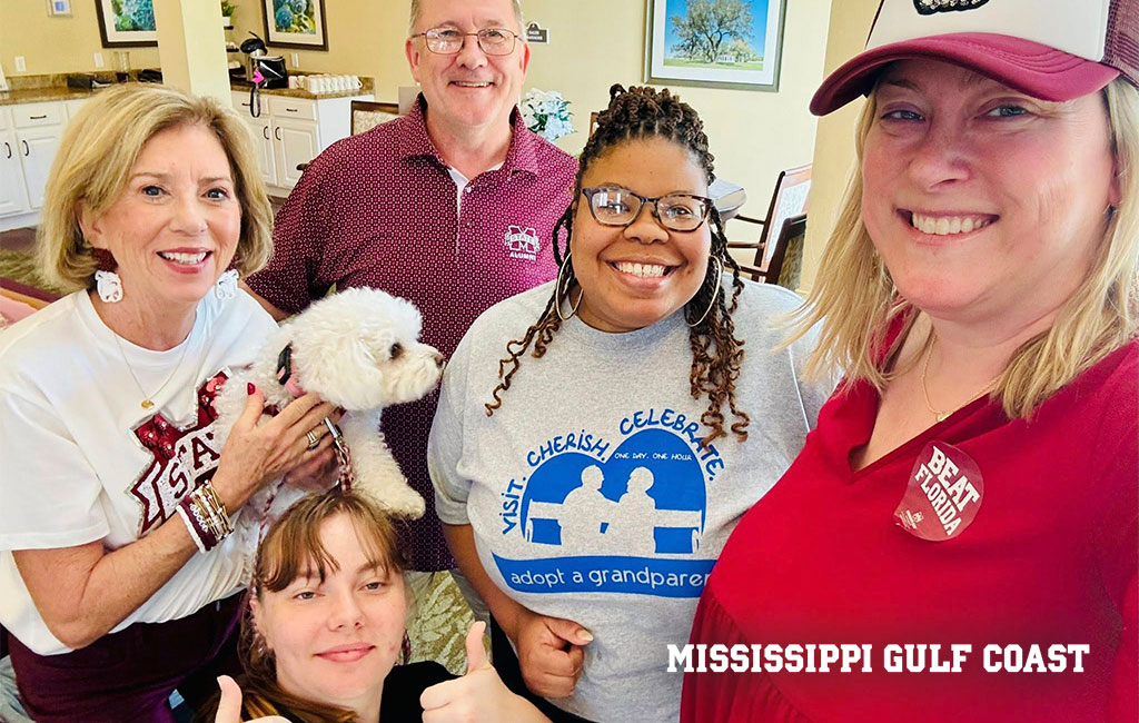 Five adults and a small white dog pose for a group photo in a brightly lit indoor setting. One person wears a “Mississippi Gulf Coast” shirt and another has a “Beat Bama” cap.