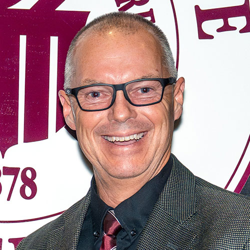 A smiling man with short gray hair and black glasses wearing a dark suit, red tie, and black shirt stands in front of a white and maroon logo backdrop.