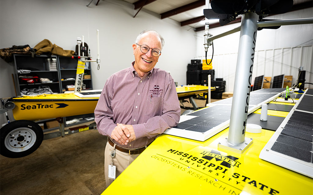 An older man in glasses and a maroon shirt stands smiling beside a yellow solar-powered research vessel in a workshop with other equipment visible.