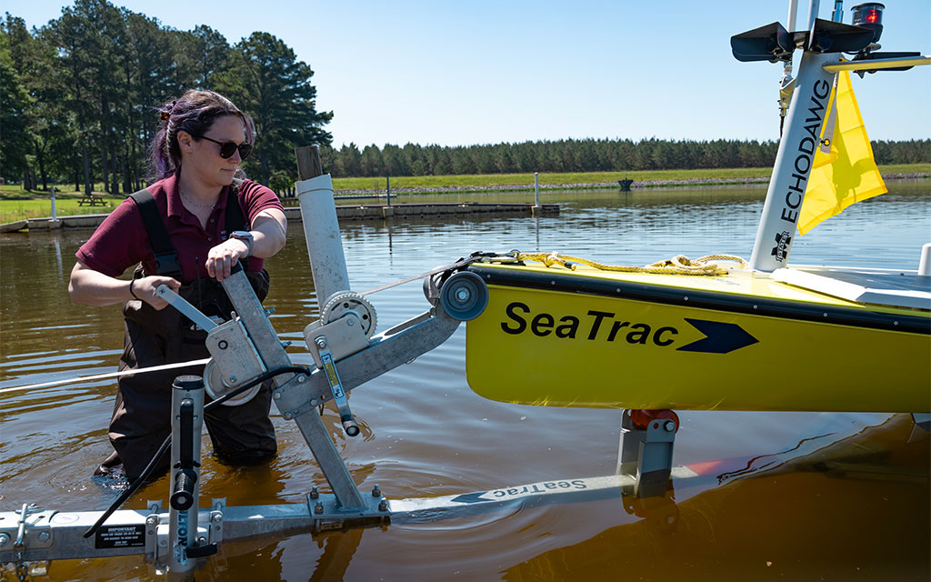 Person in waders operates a metal mechanism to guide a yellow SeaTrac boat with equipment into a shallow, calm body of water near trees.