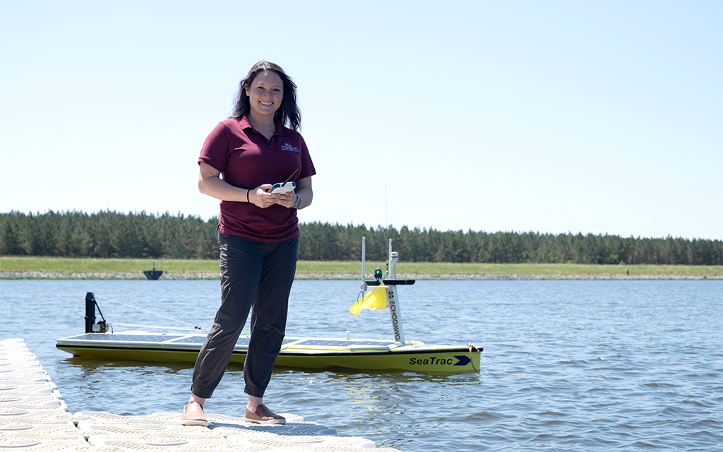 A woman stands on a dock holding a device, with a yellow SeaTrac unmanned boat on the water behind her; trees line the background under a clear sky.
