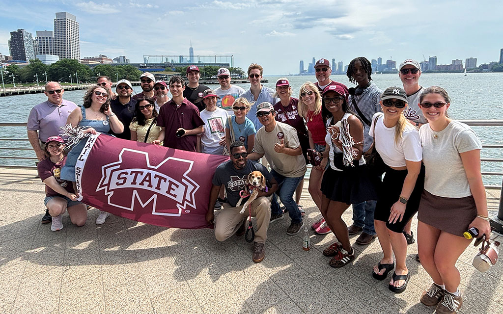 A group of people pose together outdoors by the water, holding a Mississippi State banner and wearing school apparel, with city buildings visible in the background.