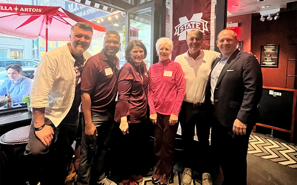 Six people pose together indoors near a Mississippi State University banner; most are wearing maroon or white clothing and name tags.