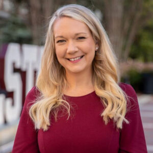 A woman with long blonde hair wearing a maroon top stands outdoors, smiling at the camera. Large white letters and greenery are visible in the blurred background.