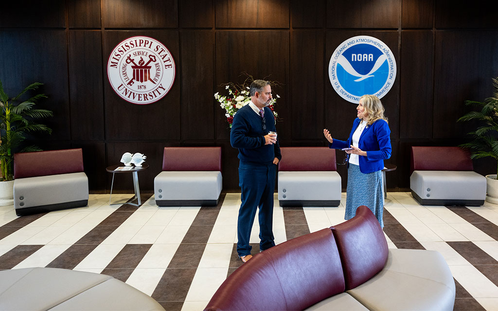 Two people stand talking in a modern lobby with Mississippi State University and NOAA logos on the wall behind them.