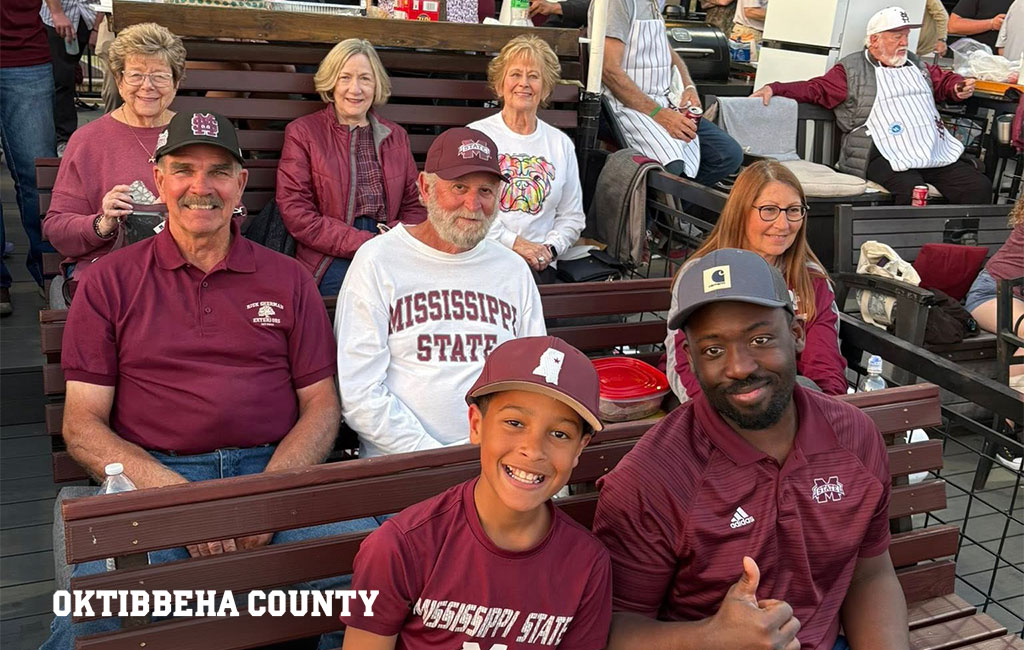 Group of people wearing Mississippi State attire sit together outdoors, smiling at the camera. A sign reading “Oktibbeha County” appears in the corner.