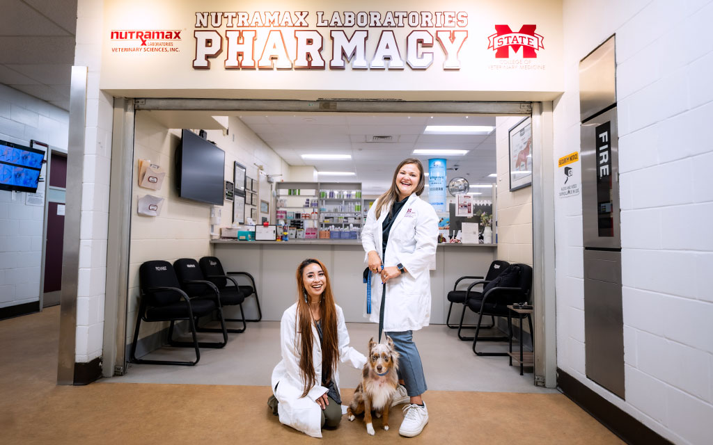 Two women in lab coats, one standing and one kneeling beside a dog, pose in front of a veterinary pharmacy labeled “Nutramax Laboratories Pharmacy.”.