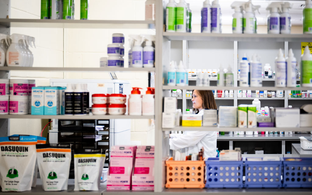 A woman in a lab coat stands behind shelves stocked with various health products, bottles, and containers in a pharmacy or laboratory setting.